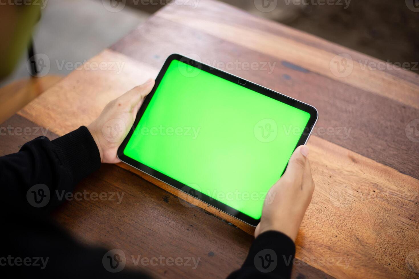 Mock up photo of a close up shot featuring a mans hand holding an iPad tablet with a green screen against the background of a wood cafe table