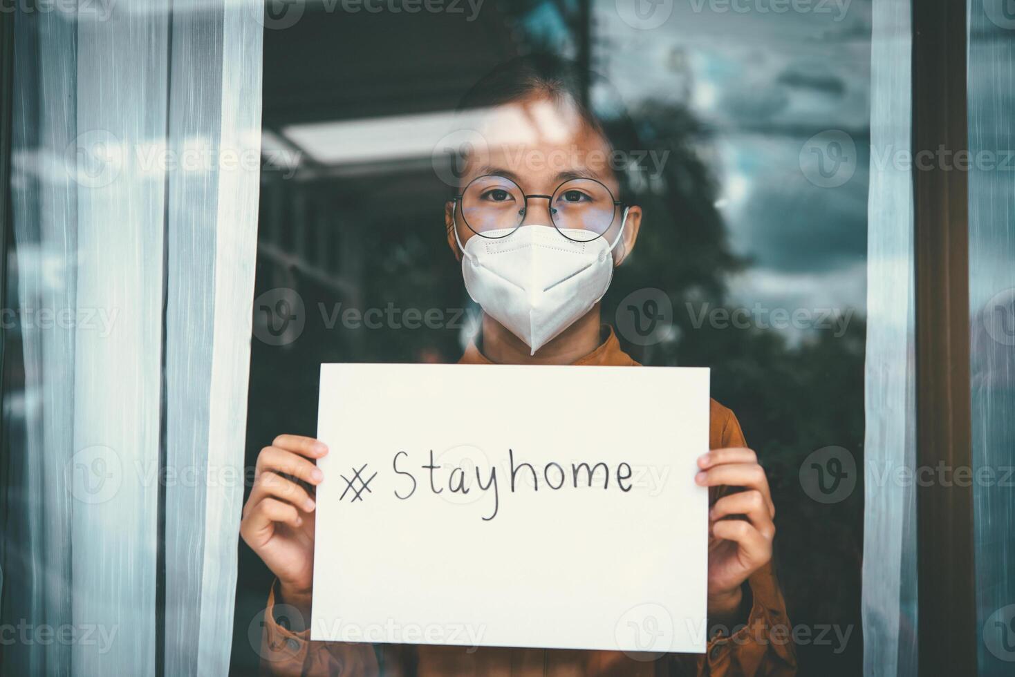 Asian girl wearing glasses, wearinglone. Concept home quarantin a mask, holding paper labeled stayhome, is bored of having to detain and treat the illness at home ae, prevention COVID-19 photo