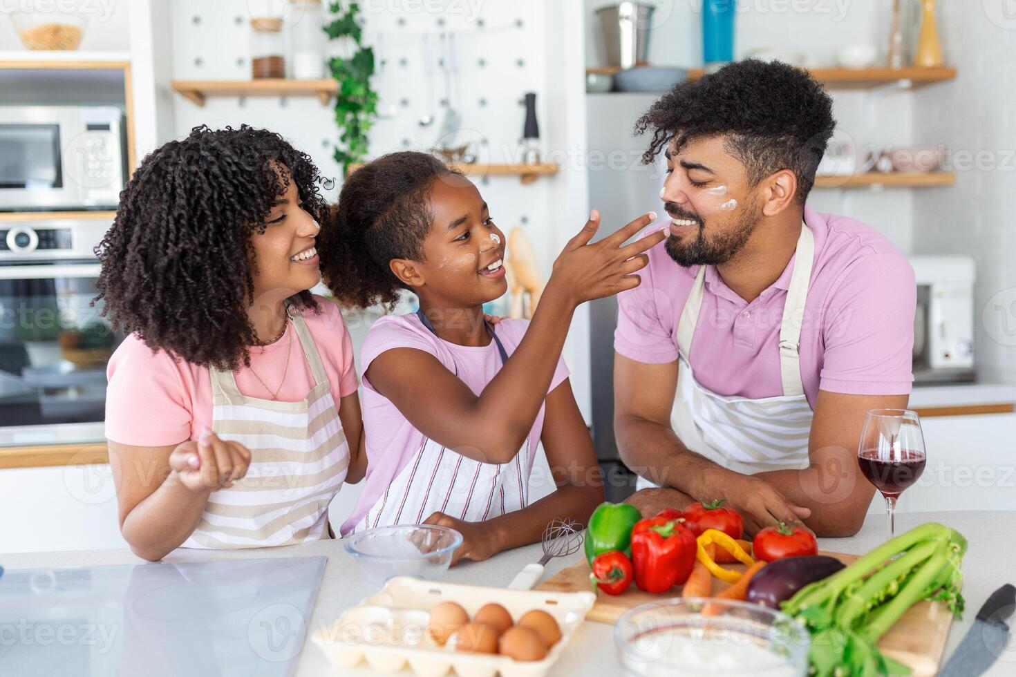 Cute little girl and her beautiful parents are smiling while cooking in kitchen at home. Happy african american family preparing healthy food together in kitchen photo