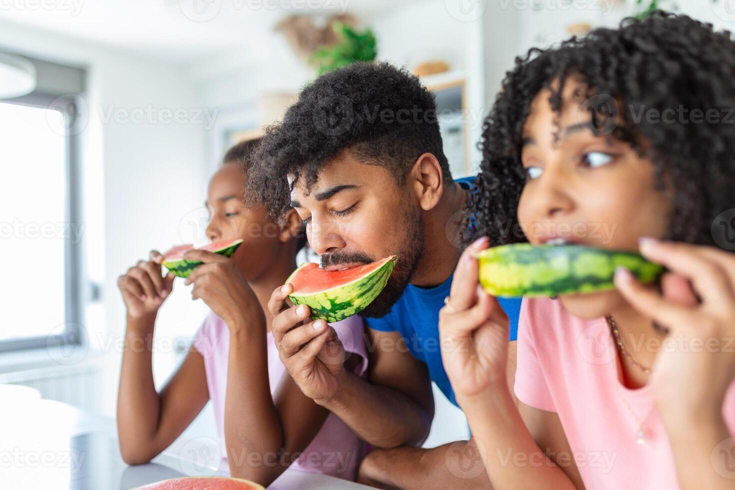 retrato de alegre joven familia tomando un mordedura de un sandía. africano americano familia en pie juntos a un cocina comiendo sandía. foto