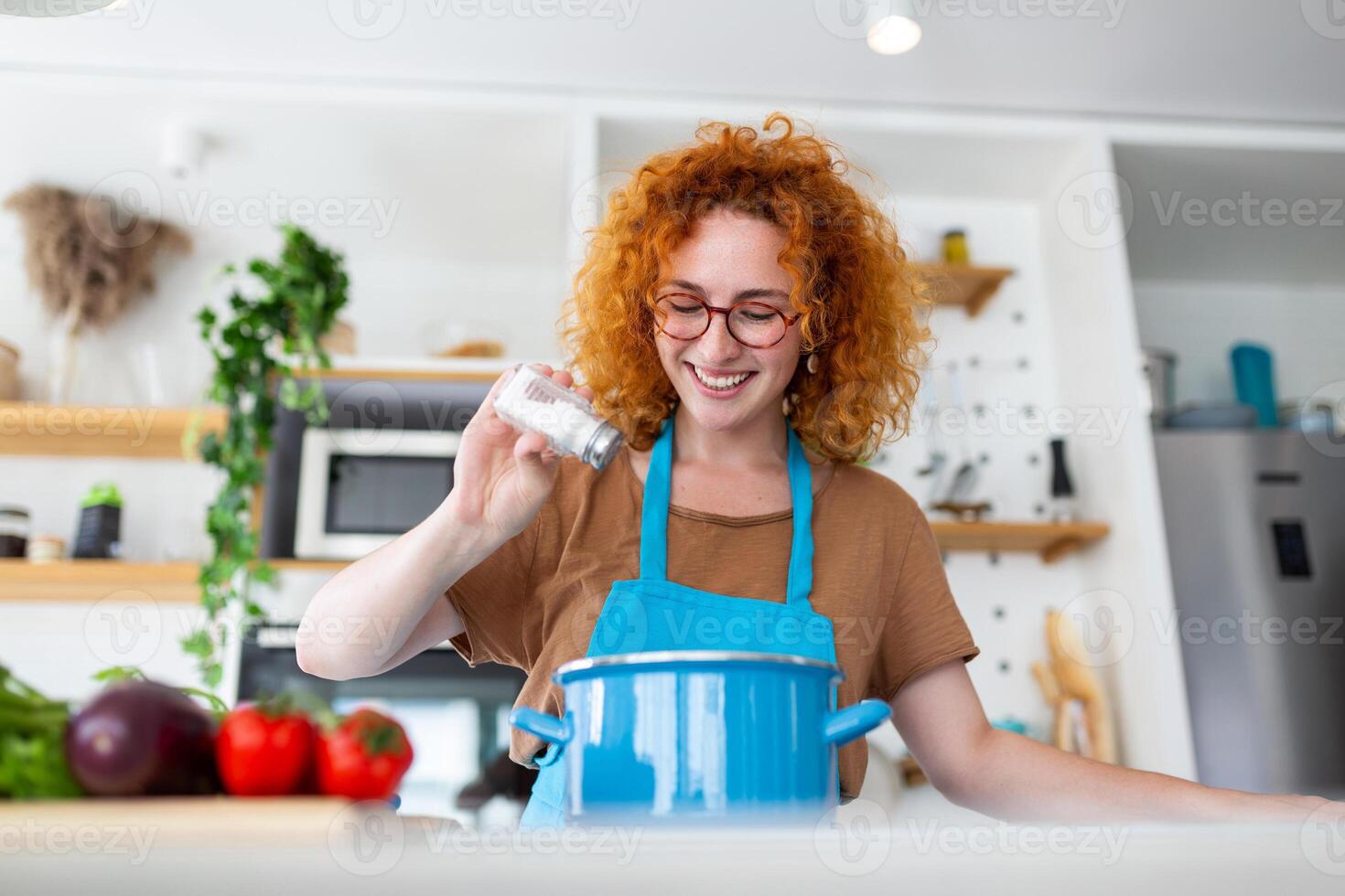 Cute young woman cooking and adding spice to meal, laughing and spending time in the kitchen photo