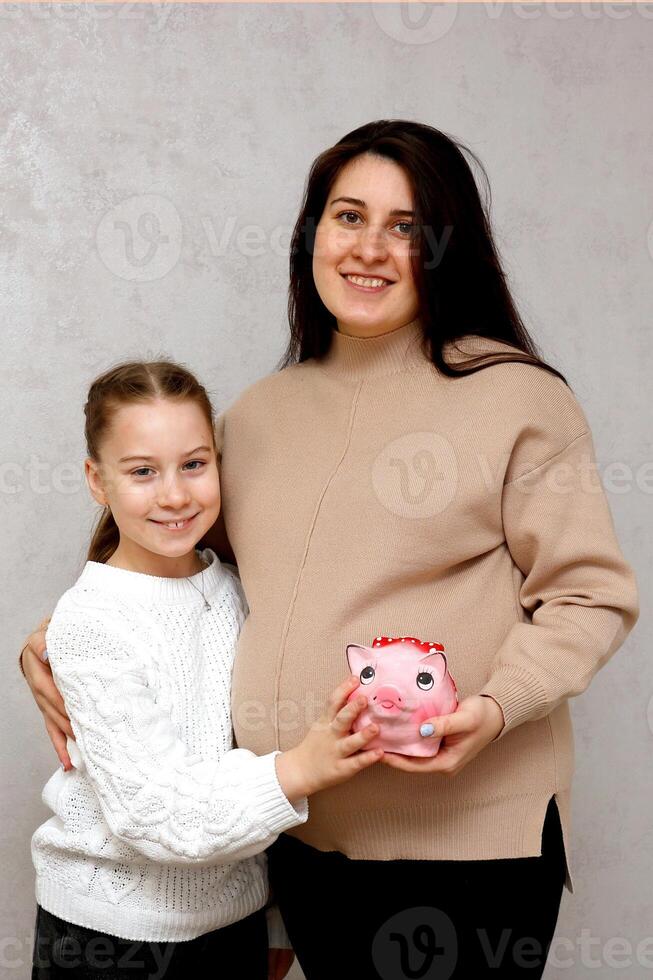 embarazada mujer con pequeño hija participación un cerdito banco en su manos foto