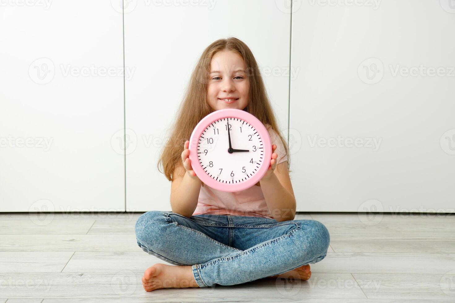 girl sits on the floor and holds a watch in her hands that shows 3 o'clock p.m. photo