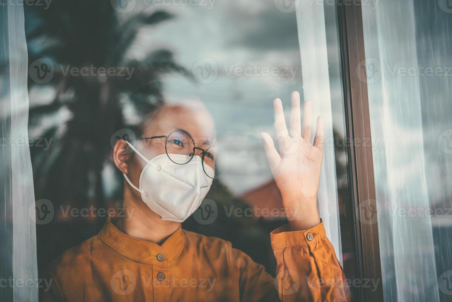 Asian girl wearing glasses, wearing a mask, looking out the window, is bored of having to detain and treat the illness at home alone. Concept home quarantine, prevention COVID-19 photo
