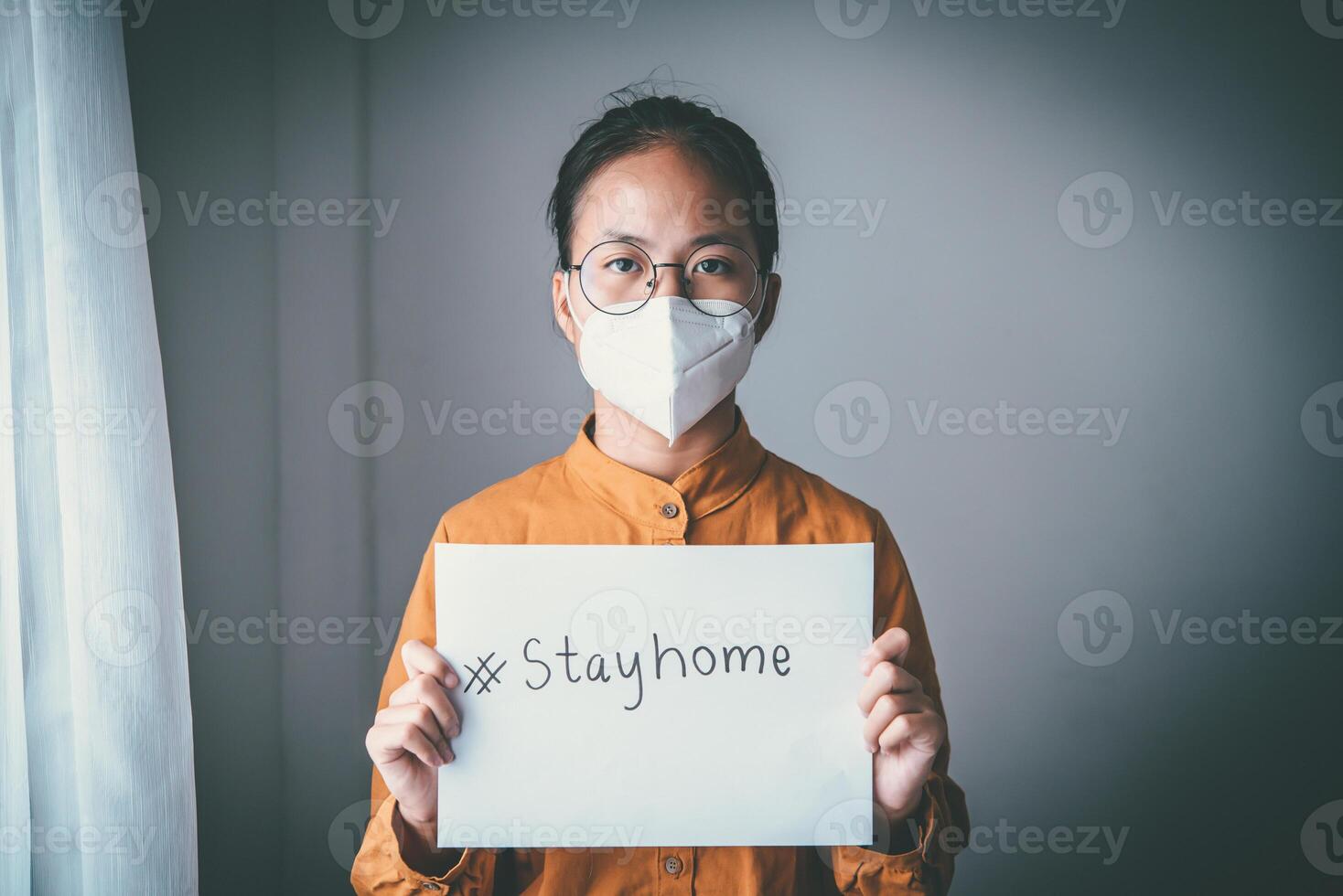 Asian girl wearing glasses, wearing a mask, holding paper labeled stayhome, is bored of having to detain and treat the illness at home alone. Concept home quarantine, prevention COVID-19 photo