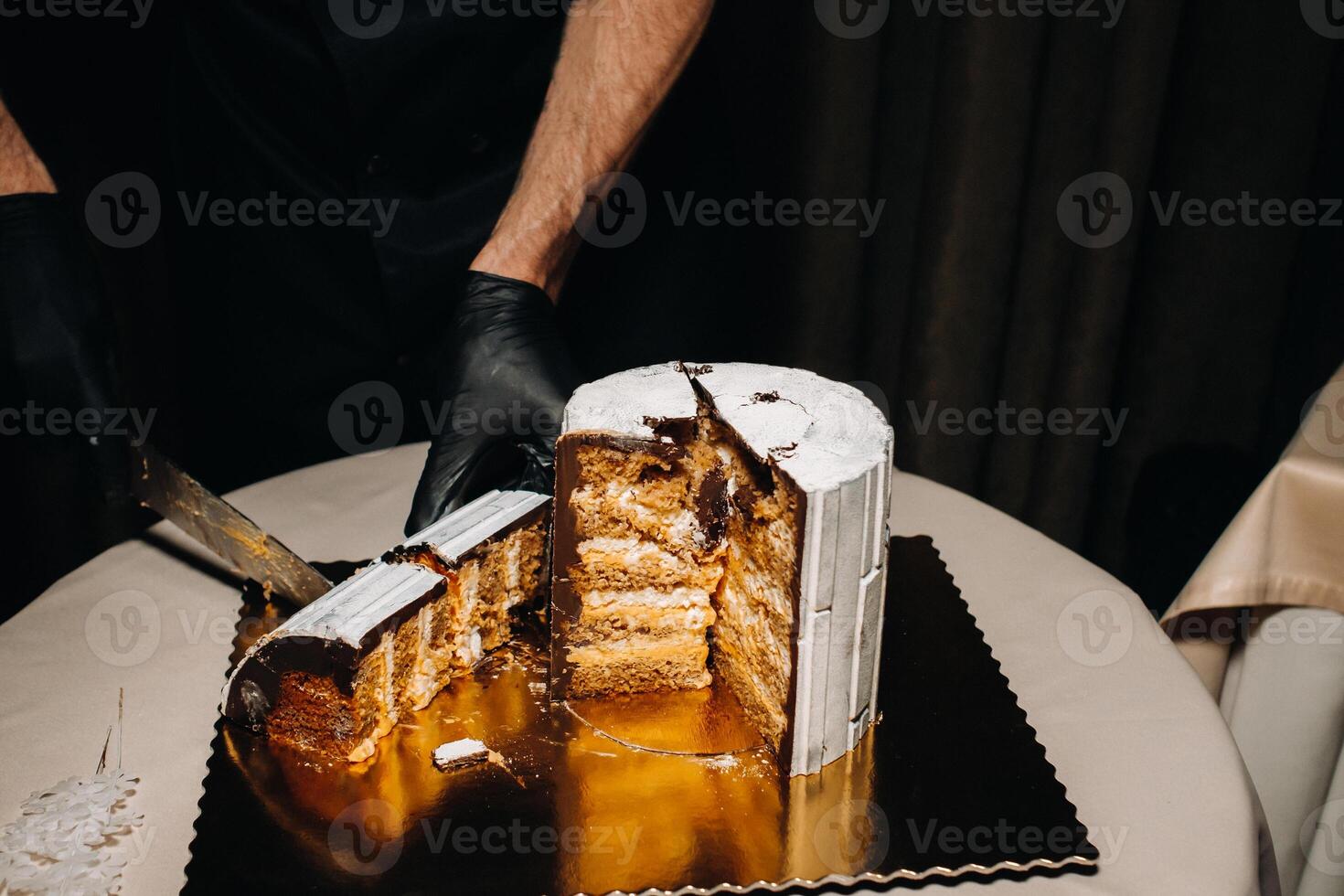 increíble pasteles un con guantes negros cocinero es rebanar un chocolate Boda pastel. el Boda pastel es delicioso dentro en un negro fondo.grande pastel en blanco chocolate. foto