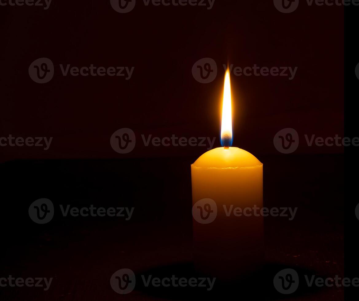 Single burning candle flame or light glowing on a big white candle on black or dark background on table in church for Christmas, funeral or memorial service with copy space. photo