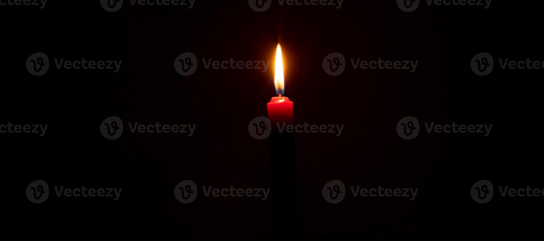 A single burning candle flame or light glowing on red candle on black or dark background on table in church for Christmas, funeral or memorial service with copy space photo