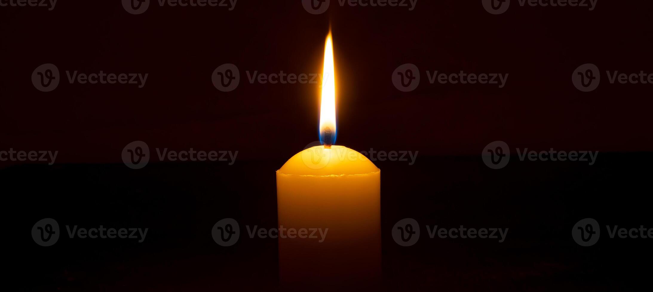 Single burning candle flame or light glowing on a big white candle on black or dark background on table in church for Christmas, funeral or memorial service with copy space. photo