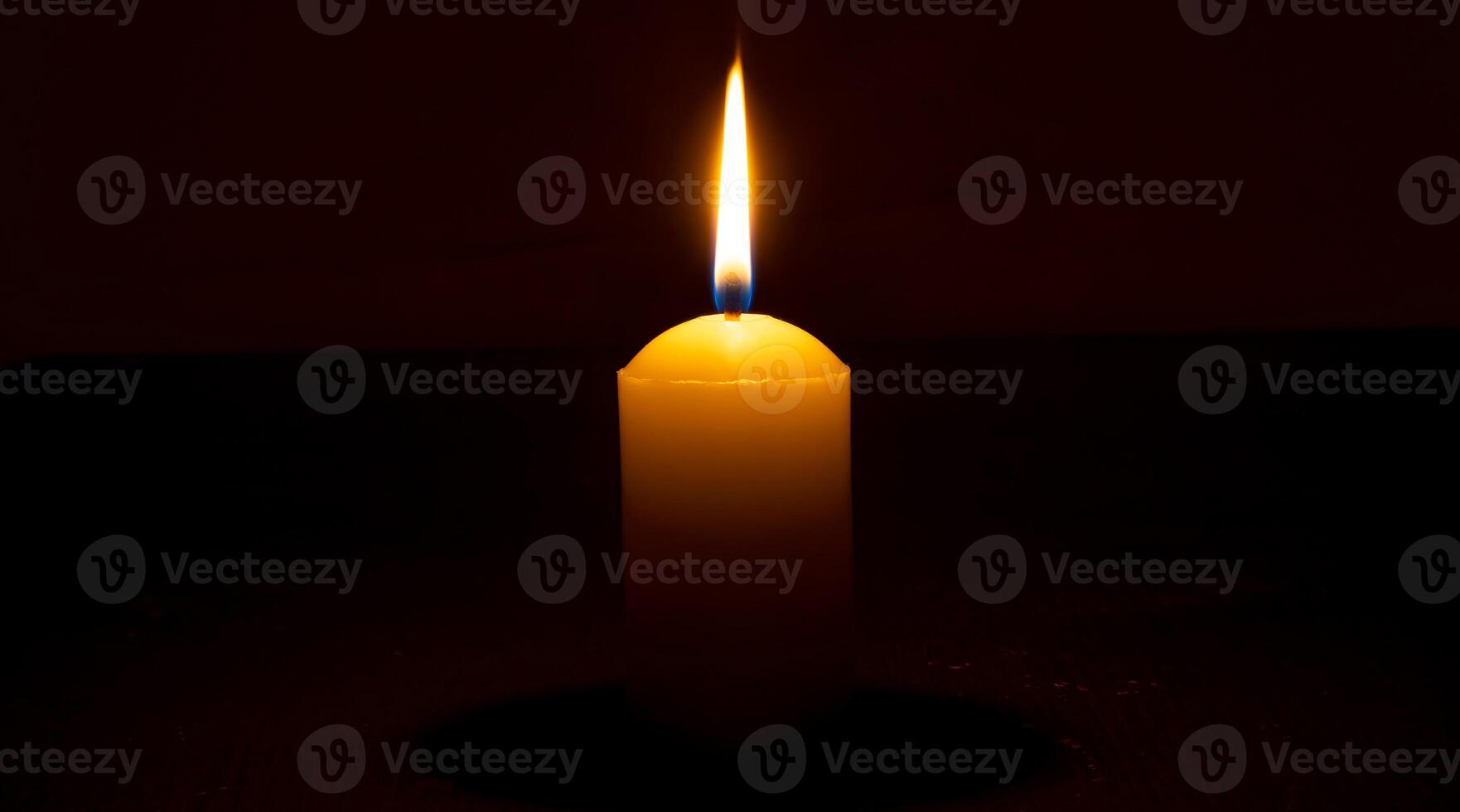 Single burning candle flame or light glowing on a big white candle on black or dark background on table in church for Christmas, funeral or memorial service with copy space. photo