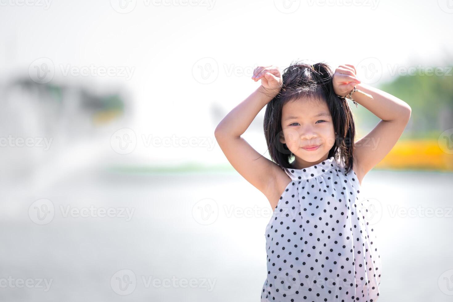 Happy Young Girl Smiling in Sunlit Park, Beaming Kid with her hands on her hair enjoys radiant, sunny day in the park, with a soft-focus fountain in the background creating a serene atmosphere. photo