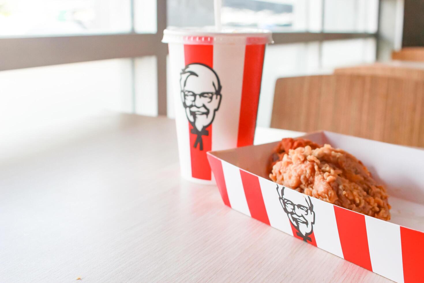 Bangkok, Thailand - January 07 2024 - Two pieces of KFC fried chicken on a paper plate and a soft drink glass with the KFC logo on a table inside a KFC restaurant. photo