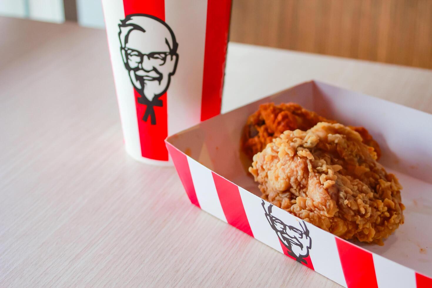 Bangkok, Thailand - January 07 2024 - Two pieces of KFC fried chicken on a paper plate and a soft drink glass with the KFC logo on a table inside a KFC restaurant. photo