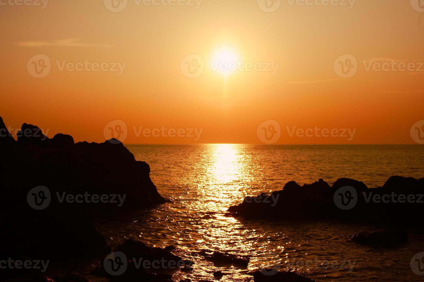 The sun is setting on the sea with rocks in the foreground, black silhouettes on the rocks, light reflecting off the sea. photo