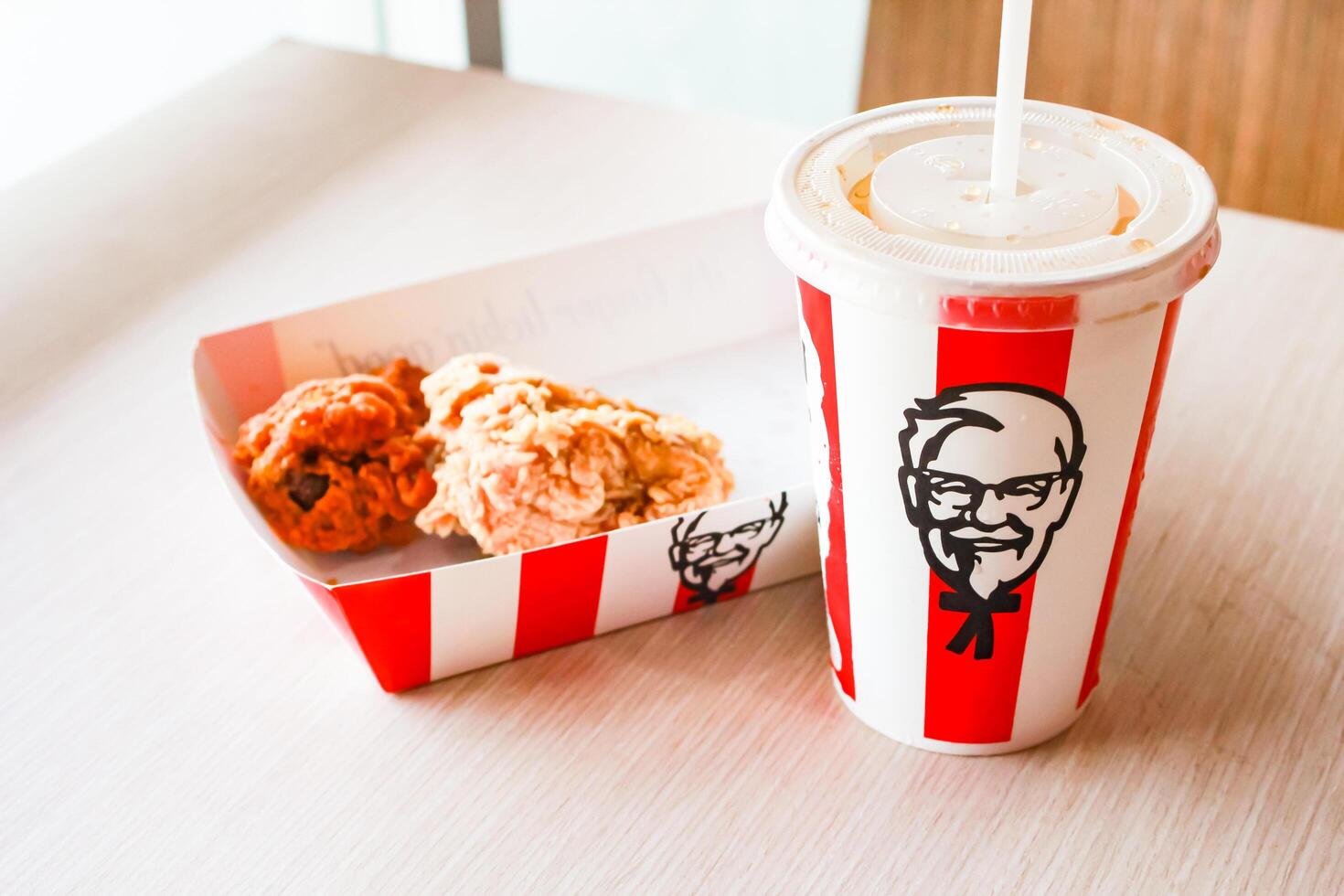Bangkok, Thailand - January 07 2024 - Two pieces of KFC fried chicken on a paper plate and a soft drink glass with the KFC logo on a table inside a KFC restaurant. photo