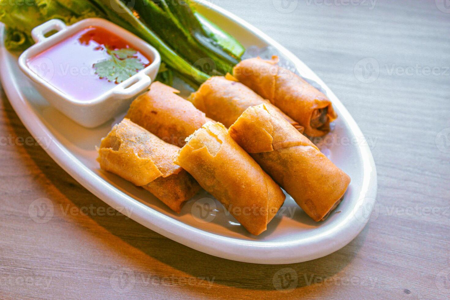 Freshly fried golden-yellow spring rolls are placed on a white plate with various vegetables and a delicious dipping sauce. photo