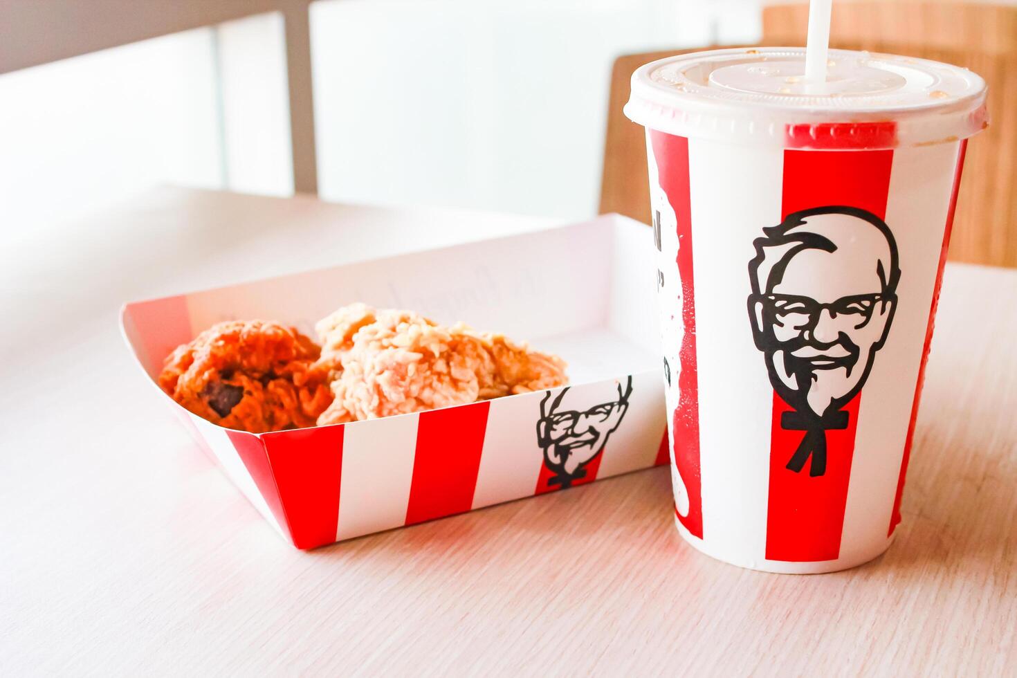 Bangkok, Thailand - January 07 2024 - Two pieces of KFC fried chicken on a paper plate and a soft drink glass with the KFC logo on a table inside a KFC restaurant. photo