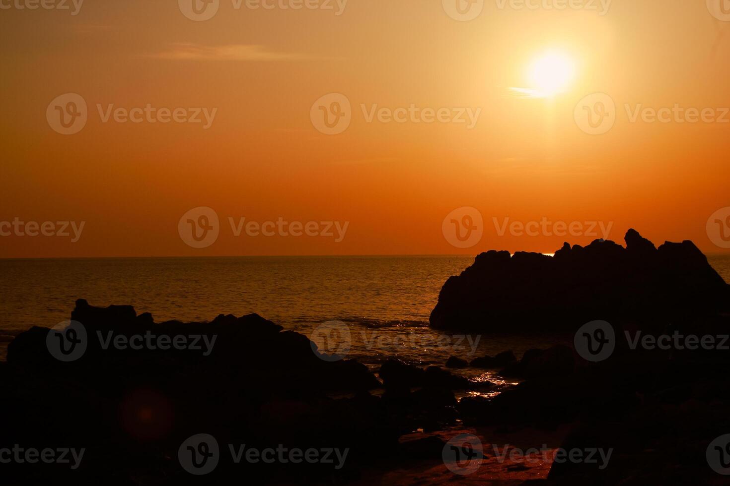 The sun is setting on the sea with rocks in the foreground, black silhouettes on the rocks, light reflecting off the sea. photo