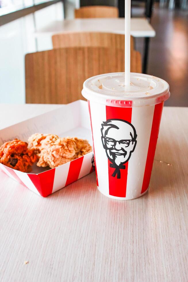 Bangkok, Thailand - January 07 2024 - Two pieces of KFC fried chicken on a paper plate and a soft drink glass with the KFC logo on a table inside a KFC restaurant. photo