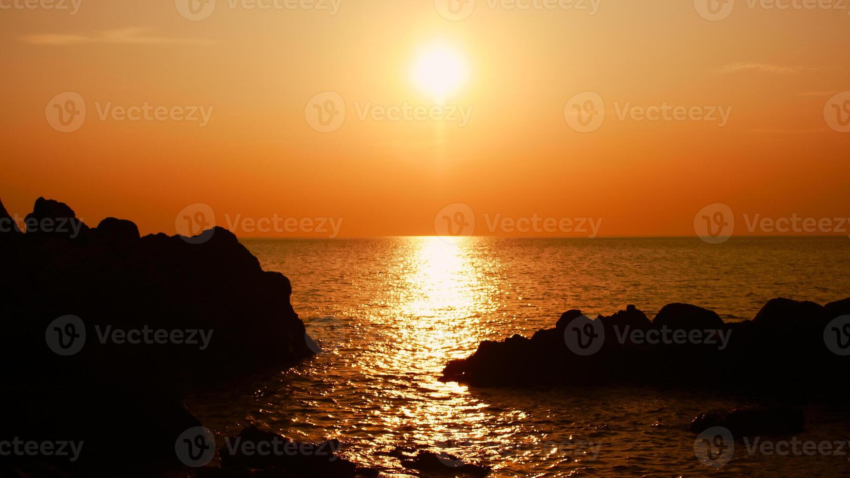 The sun is setting on the sea with rocks in the foreground, black silhouettes on the rocks, light reflecting off the sea. photo