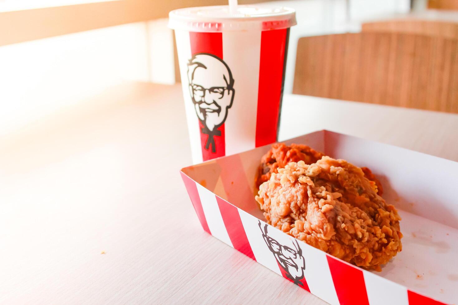 Bangkok, Thailand - January 07 2024 - Two pieces of KFC fried chicken on a paper plate and a soft drink glass with the KFC logo on a table inside a KFC restaurant. photo