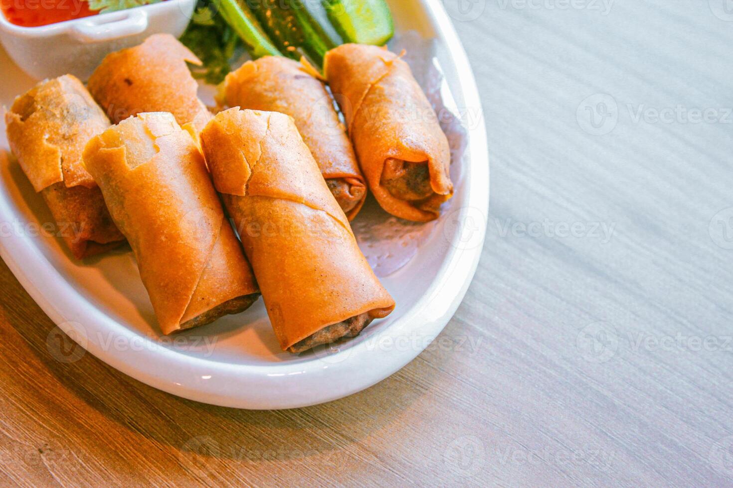 Freshly fried golden-yellow spring rolls are placed on a white plate with various vegetables and a delicious dipping sauce. photo