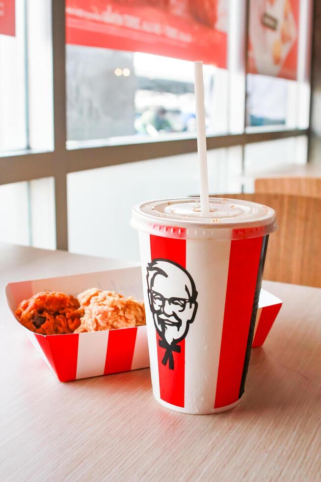 Bangkok, Thailand - January 07 2024 - Two pieces of KFC fried chicken on a paper plate and a soft drink glass with the KFC logo on a table inside a KFC restaurant. photo