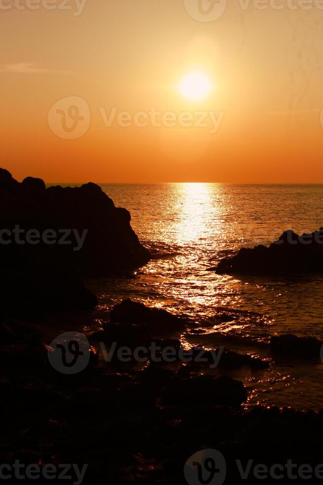 The sun is setting on the sea with rocks in the foreground, black silhouettes on the rocks, light reflecting off the sea. photo