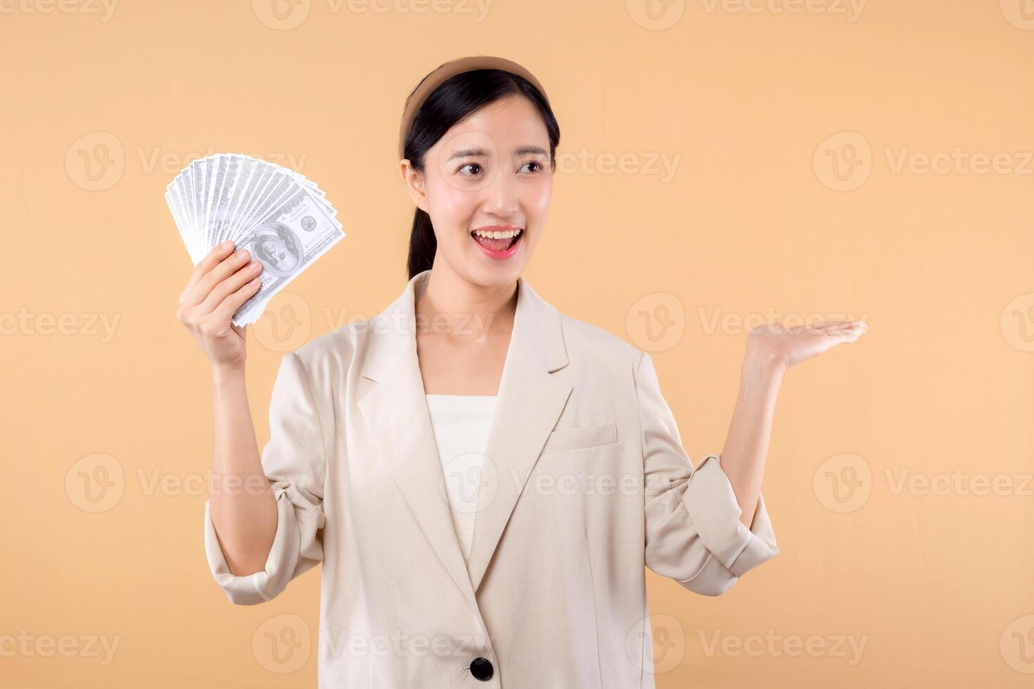 portrait of happy successful confident young asian business woman wearing white jacket holding cash money dollars standing over beige background. millionaire business, shopping concept. photo