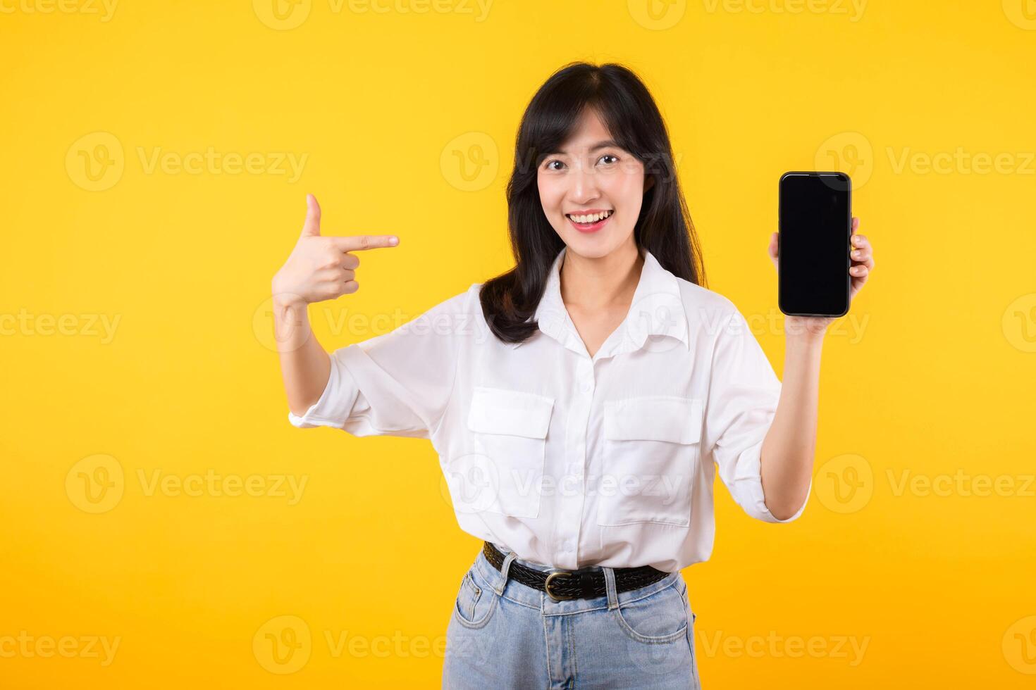 Portrait beautiful young asian woman happy smile wearing white shirt and denim plants showing pointing finger hand gesture to smartphone isolated on yellow studio background. app smartphone concept. photo