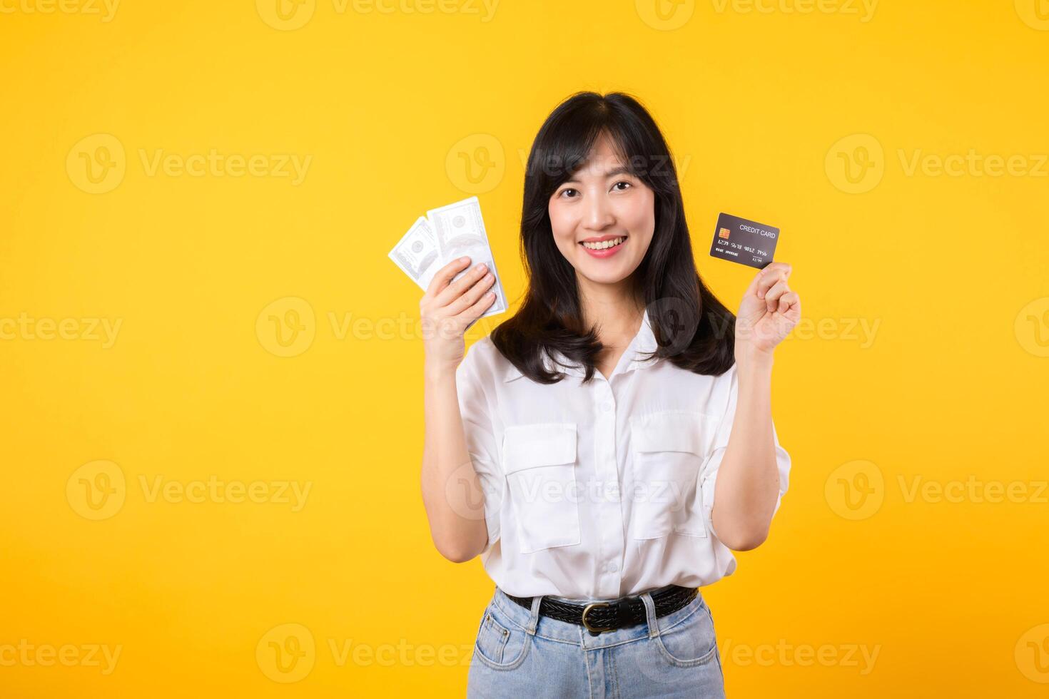 happy successful confident young asian woman happy smile wearing white shirt and denim jean holding cash money and credit card standing over yellow background. millionaire business, shopping concept. photo