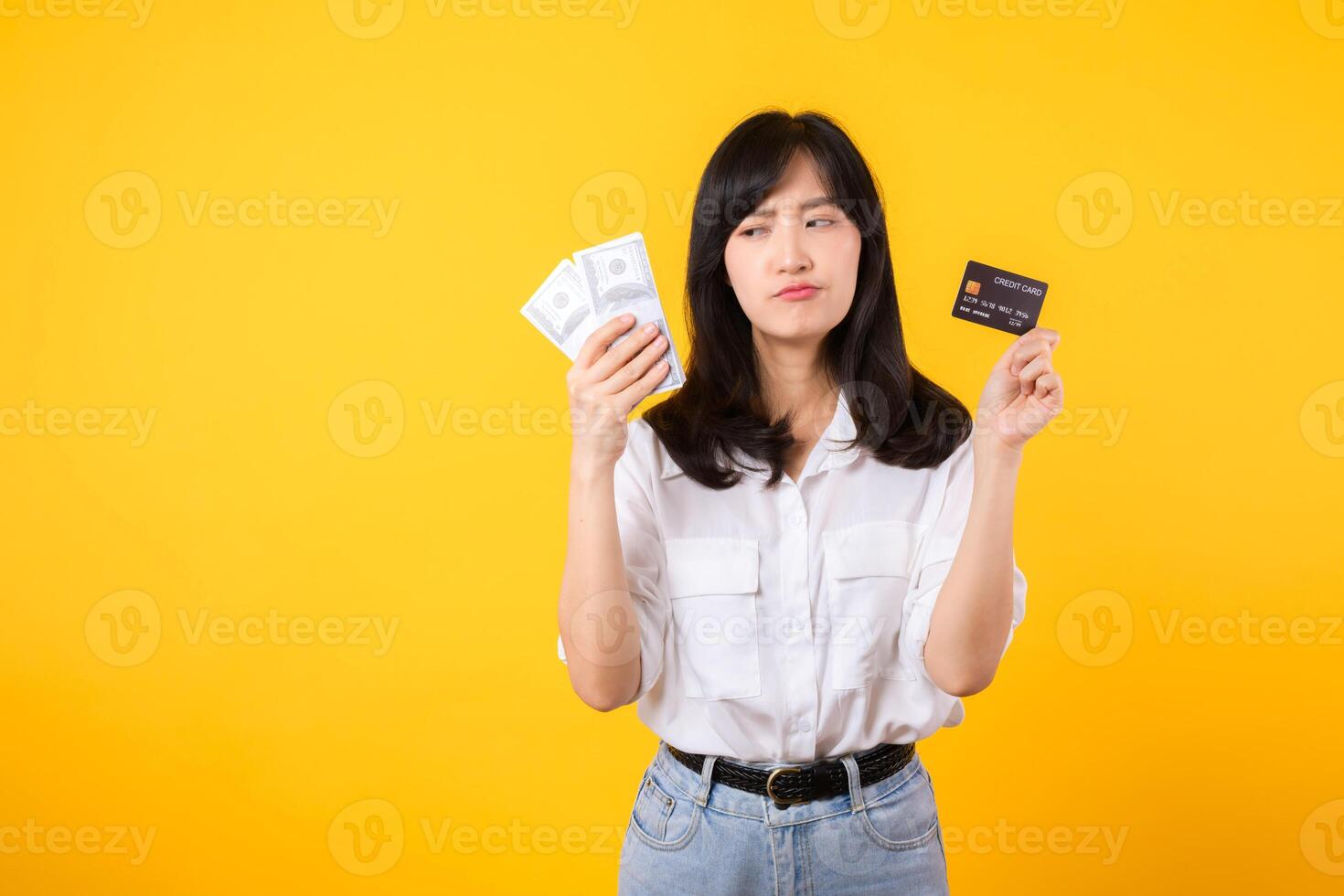 happy successful confident young asian woman happy smile wearing white shirt and denim jean holding cash money and credit card standing over yellow background. millionaire business, shopping concept. photo