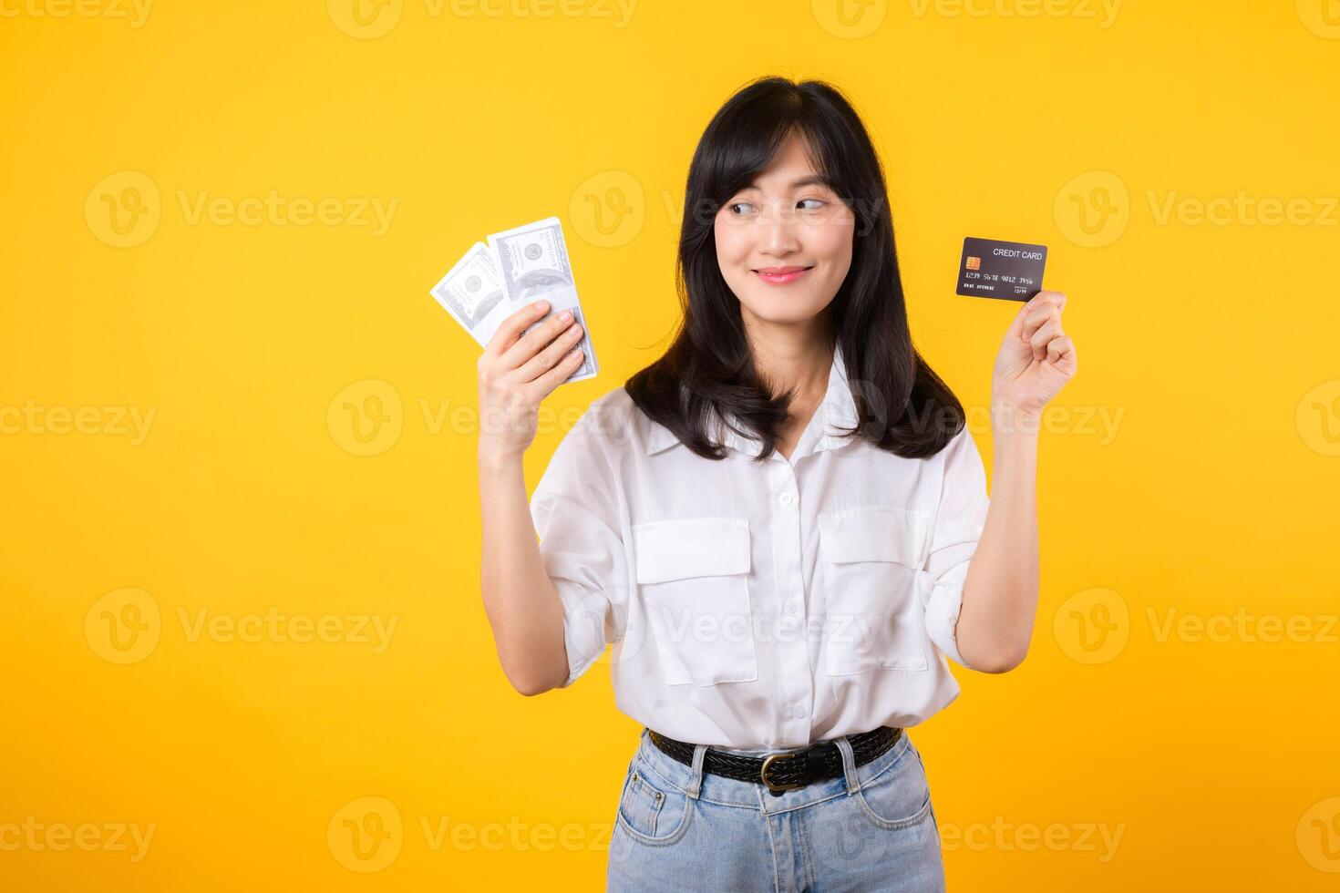 happy successful confident young asian woman happy smile wearing white shirt and denim jean holding cash money and credit card standing over yellow background. millionaire business, shopping concept. photo