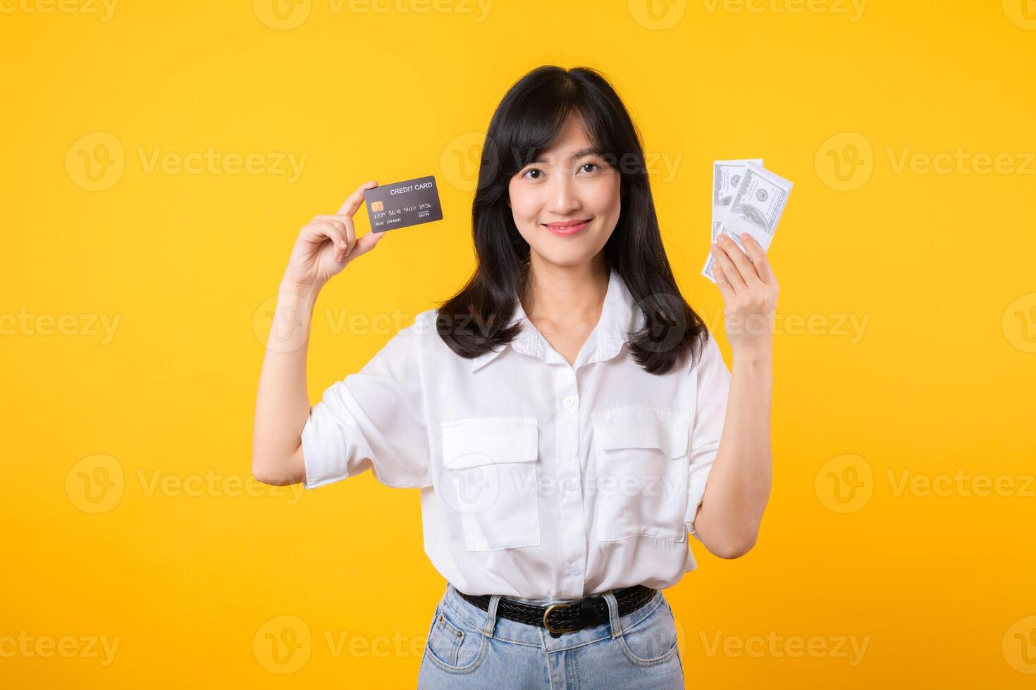 happy successful confident young asian woman happy smile wearing white shirt and denim jean holding cash money and credit card standing over yellow background. millionaire business, shopping concept. photo