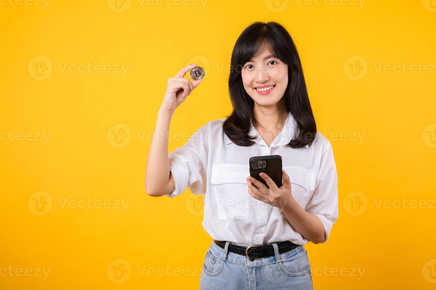 Stunning portrait of an Asian young woman holding digital currency and smartphone on yellow studio background. Explore the concept of crypto digital currency. photo
