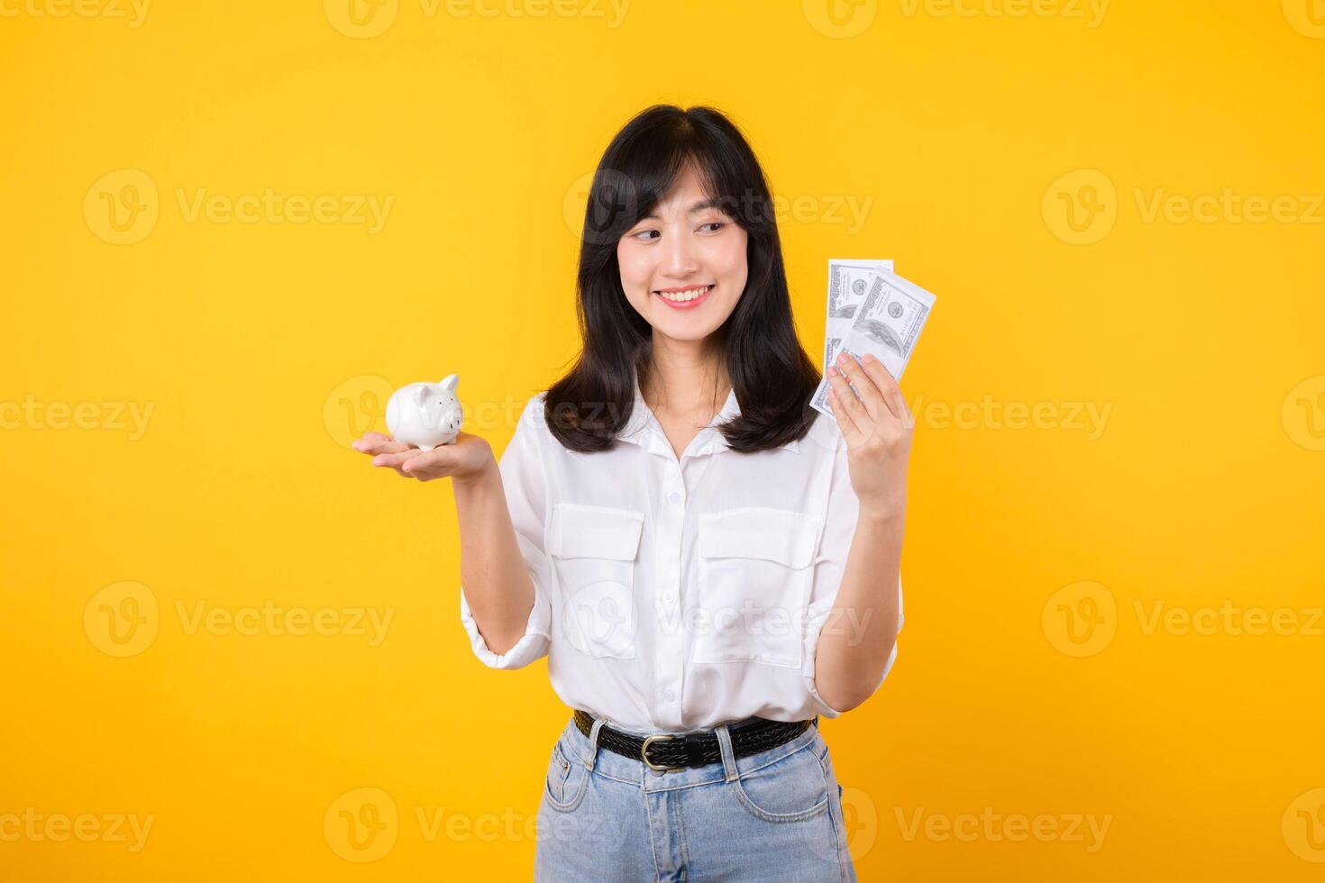 Portrait beautiful young business asian woman wearing white shirt and denim jean with a lot of cash money and piggy bank isolated on yellow background. Wealth money saving, finance investment concept. photo