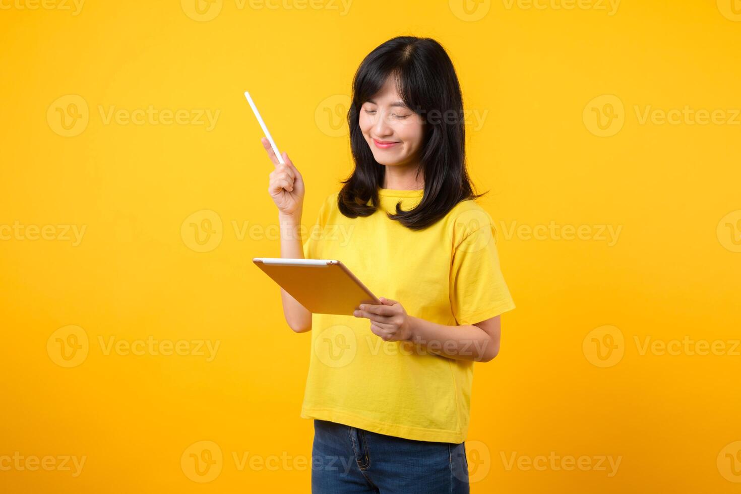young Asian woman wearing yellow t-shirt and jeans showing happy smile while using digital tablet, displaying thoughtful expression and creative idea. education technology innovative thinking concept. photo