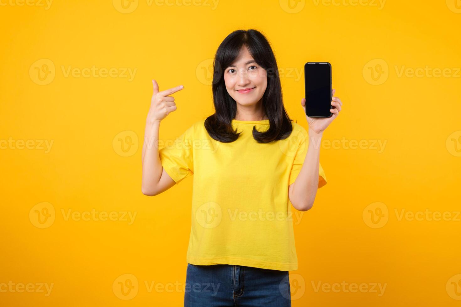 Capture joy and excitement of new applications with cheerful portrait young Asian woman wearing yellow t-shirt and denim jeans points finger towards smartphone. concept of app and digital innovation. photo
