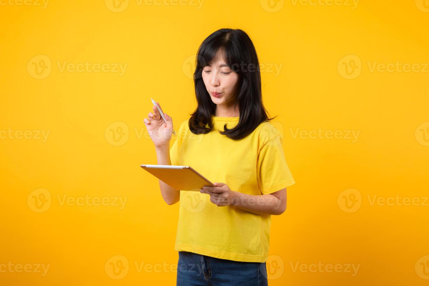 young Asian woman wearing yellow t-shirt and jeans showing happy smile while using digital tablet, displaying thoughtful expression and creative idea. education technology innovative thinking concept. photo