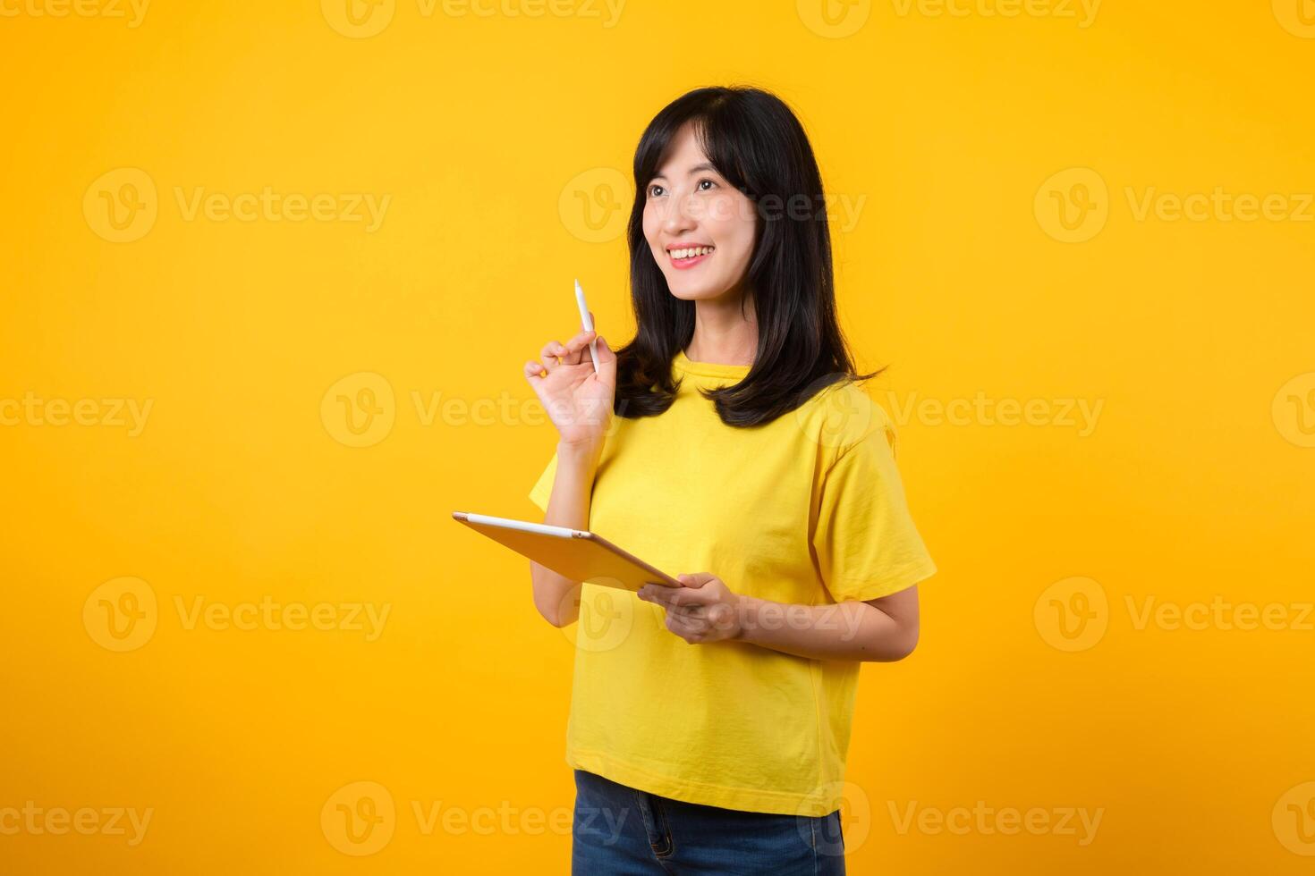 young Asian woman wearing yellow t-shirt and jeans showing happy smile while using digital tablet, displaying thoughtful expression and creative idea. education technology innovative thinking concept. photo