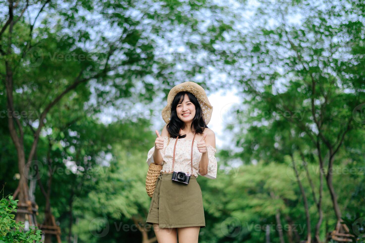 retrato de asiático joven mujer viajero con Costura sombrero y cesta y un cámara en verde público parque naturaleza antecedentes. viaje viaje estilo de vida, mundo viaje explorador o Asia verano turismo concepto. foto