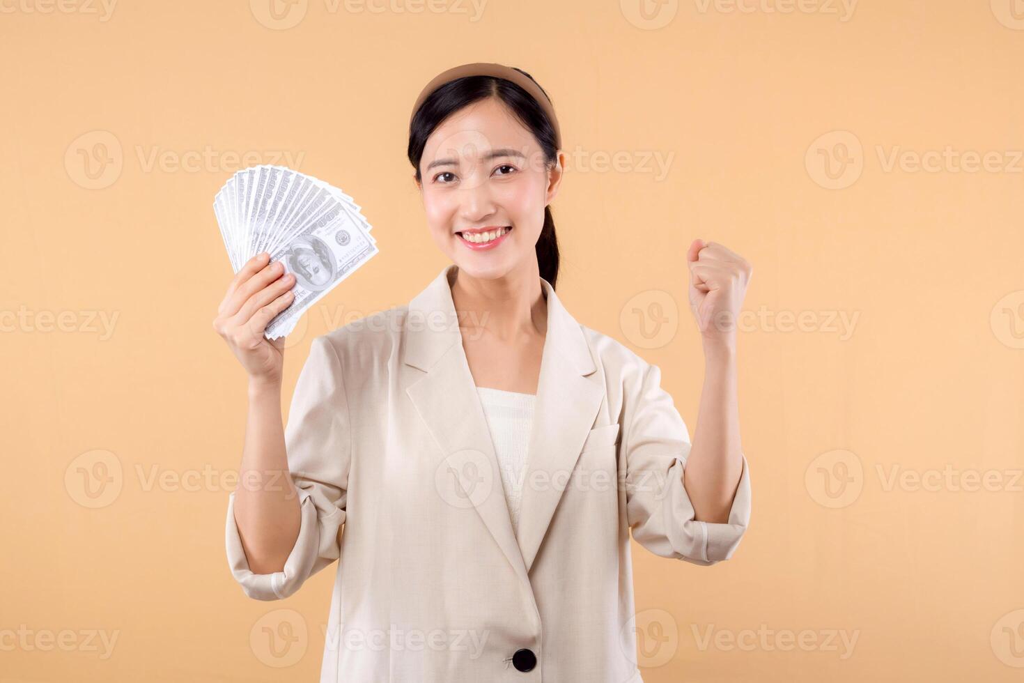 portrait of happy successful confident young asian business woman wearing white jacket holding cash money dollars standing over beige background. millionaire business, shopping concept. photo