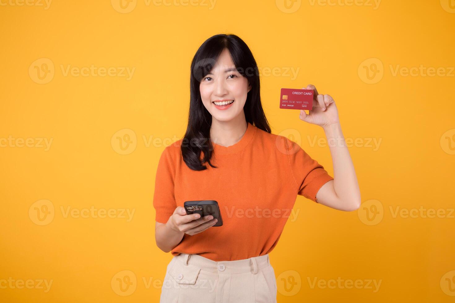 cheerful young asian woman 30s wearing orange shirt holding credit card and smartphone isolated on yellow background. symbolizing seamless digital transactions. financial freedom, convenience concept. photo