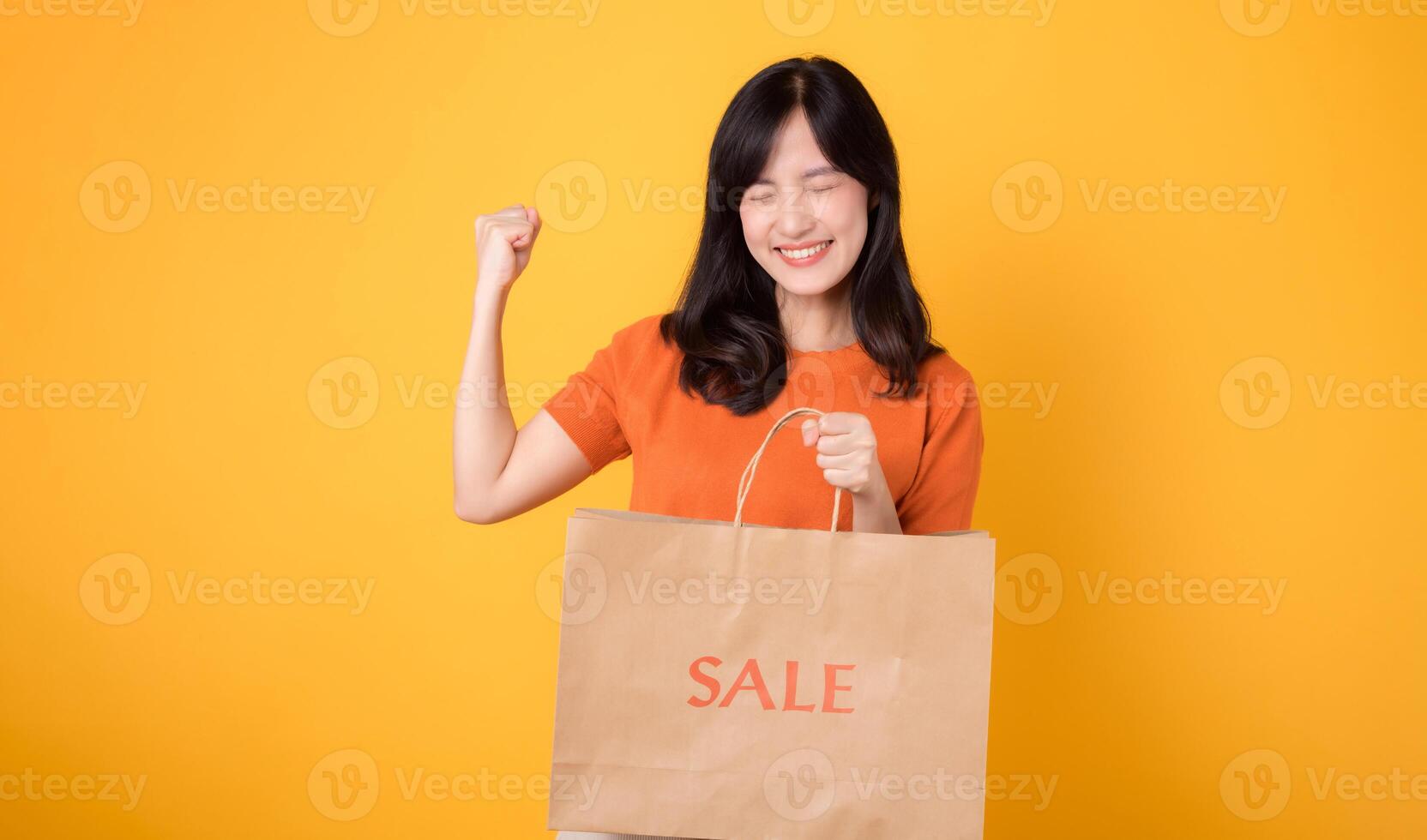 Cheerful Asian 30s woman wearing orange shirt showcases happy smile while proudly holding sale paper bag and raising fist up triumphant gesture against yellow background. Shopping, best deal concept. photo