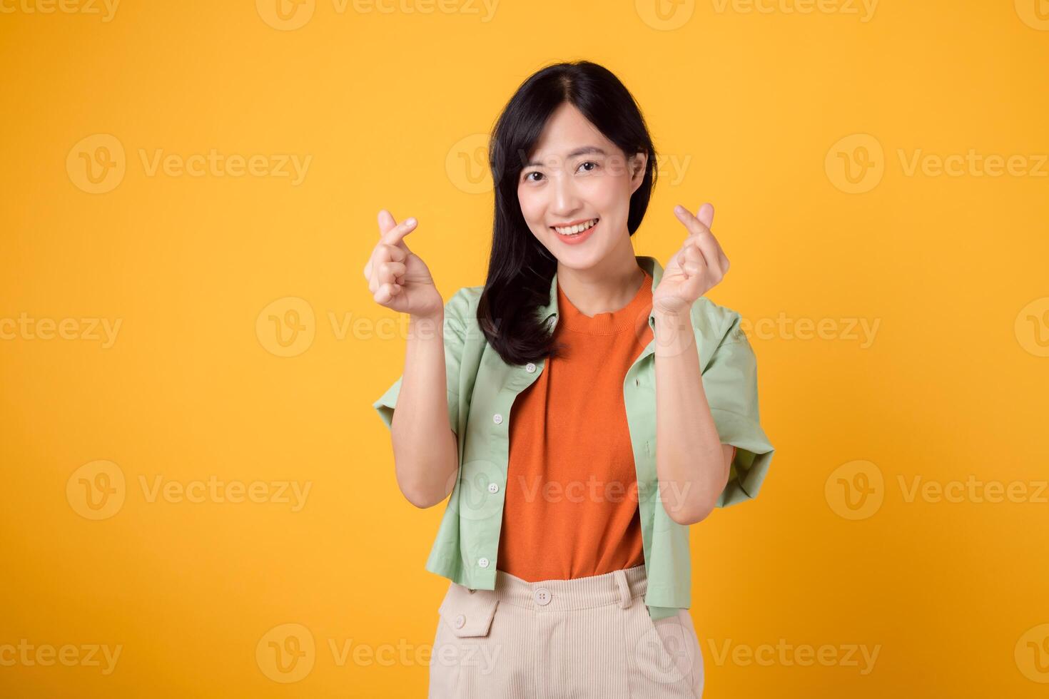 Cheerful Asian woman 30s wearing orange shirt showcases a mini heart hand sign gesture isolated on yellow background, symbolizing affection and positivity. happiness and spreading love concept. photo