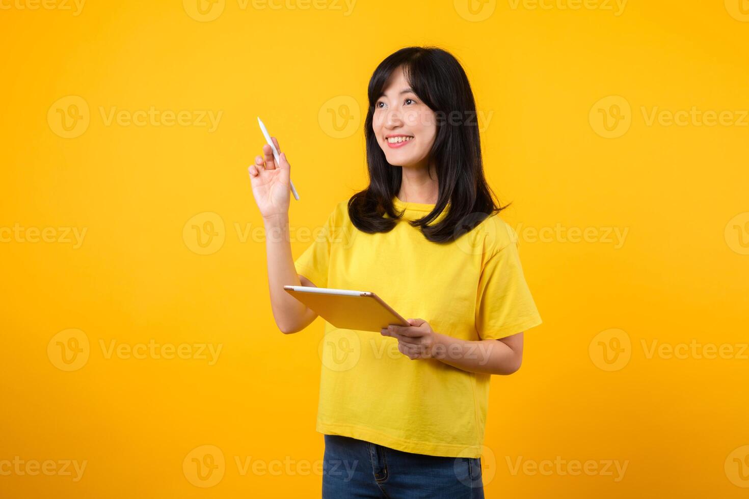 young Asian woman wearing yellow t-shirt and jeans showing happy smile while using digital tablet, displaying thoughtful expression and creative idea. education technology innovative thinking concept. photo