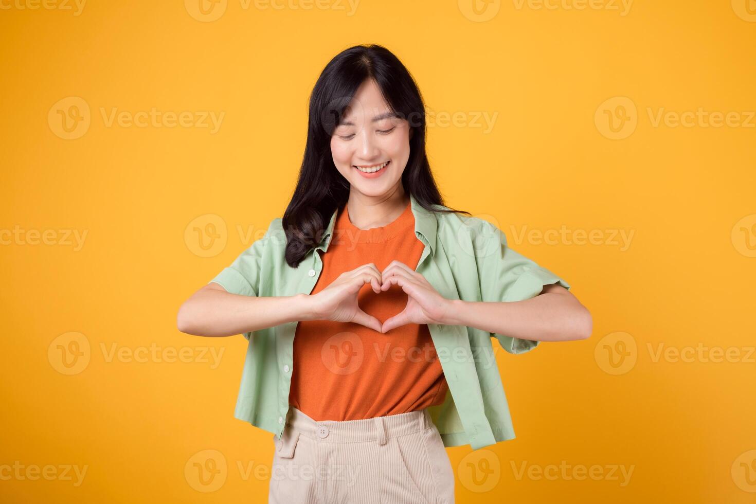 portrait grateful joy young asian woman making heart hands shape to her chest isolated on yellow background. expression peaceful, thankful emotion happiness love positive feeling. photo