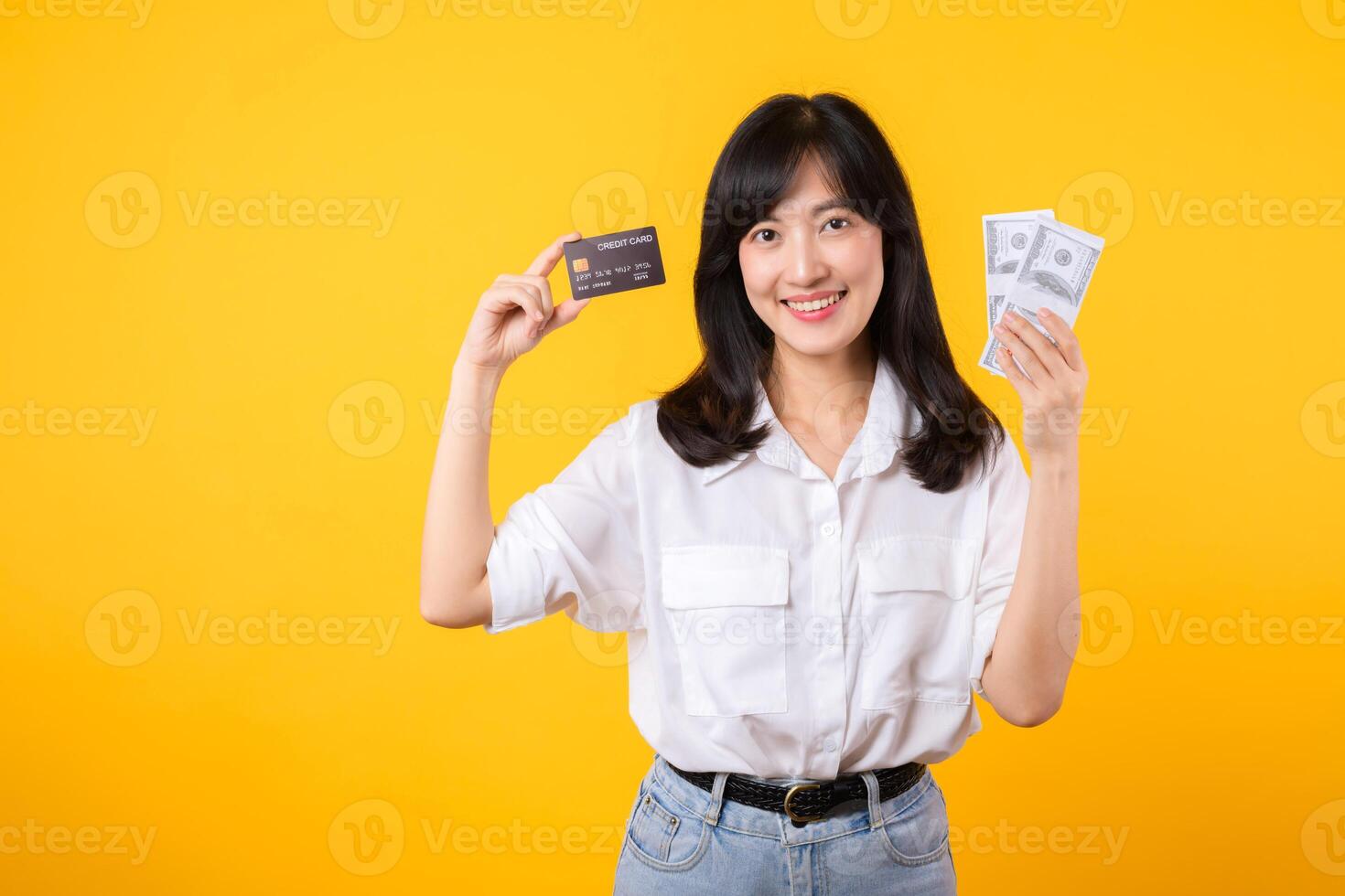 happy successful confident young asian woman happy smile wearing white shirt and denim jean holding cash money and credit card standing over yellow background. millionaire business, shopping concept. photo