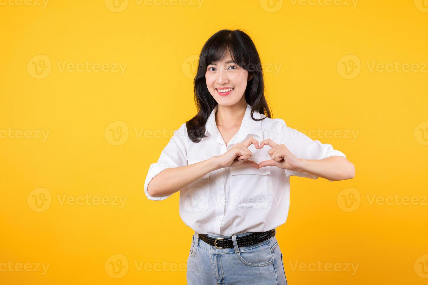 Young female healthcare. Asian woman wearing white shirt feels happy and romantic shapes heart gesture expresses tender feeling poses isolated on yellow background. People affection and care concept. photo