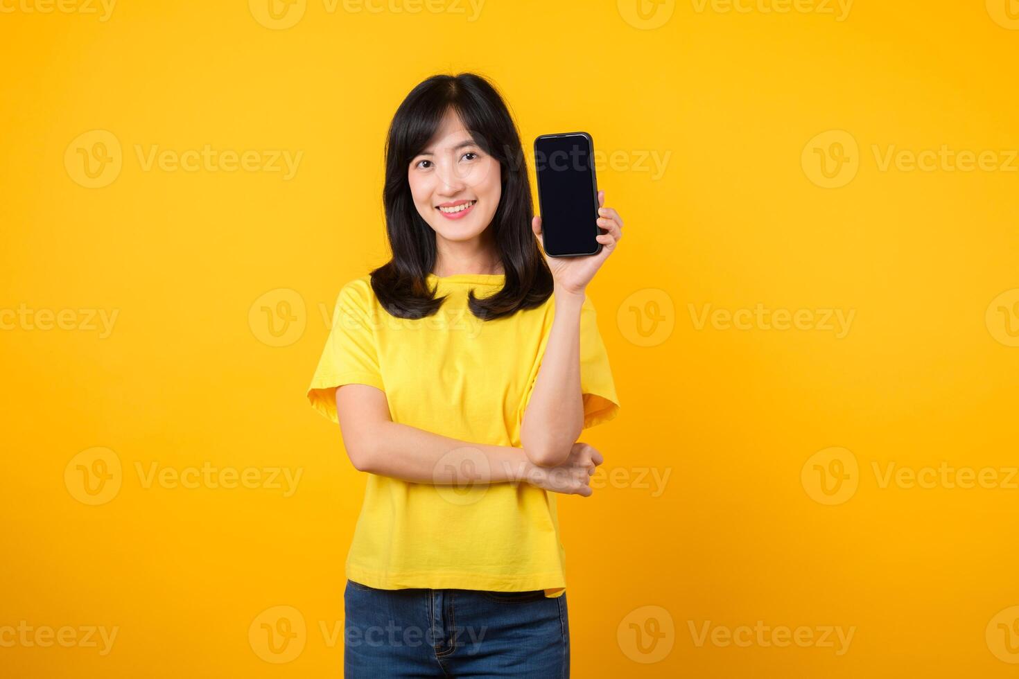 Experience joy with cheerful portrait, young Asian woman wearing yellow t-shirt and denim jeans showcases happy smile while showing smartphone screen. concept of recommended applications. photo